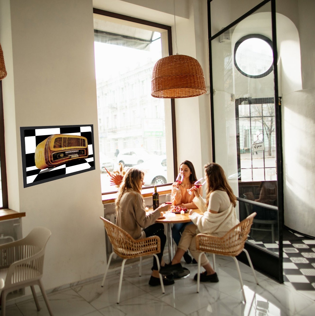 An impressive drawing of an antique radio in the style of the 1970s     
 A black frame, hanging in a small restaurant with a black and white floor. At the table three women are drinking coffee and talking